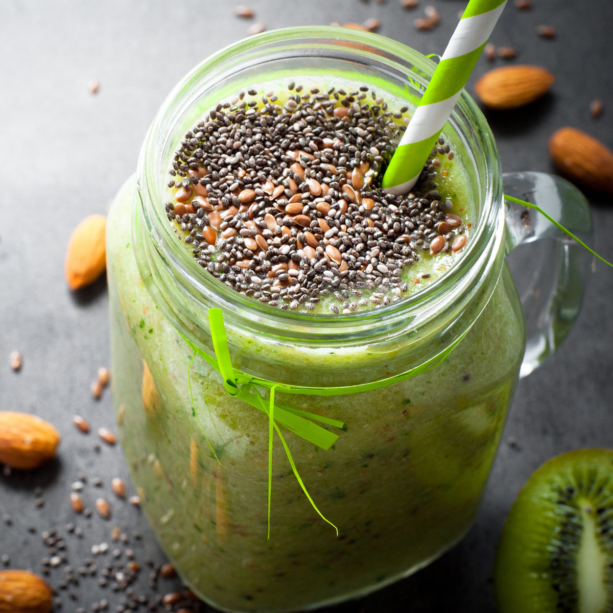 A green smoothie topped with seeds is served in a glass jar with fresh green leaves and fruit nearby, showing a drink idea using The Herb Shed Organic Linseed (Flaxseed).