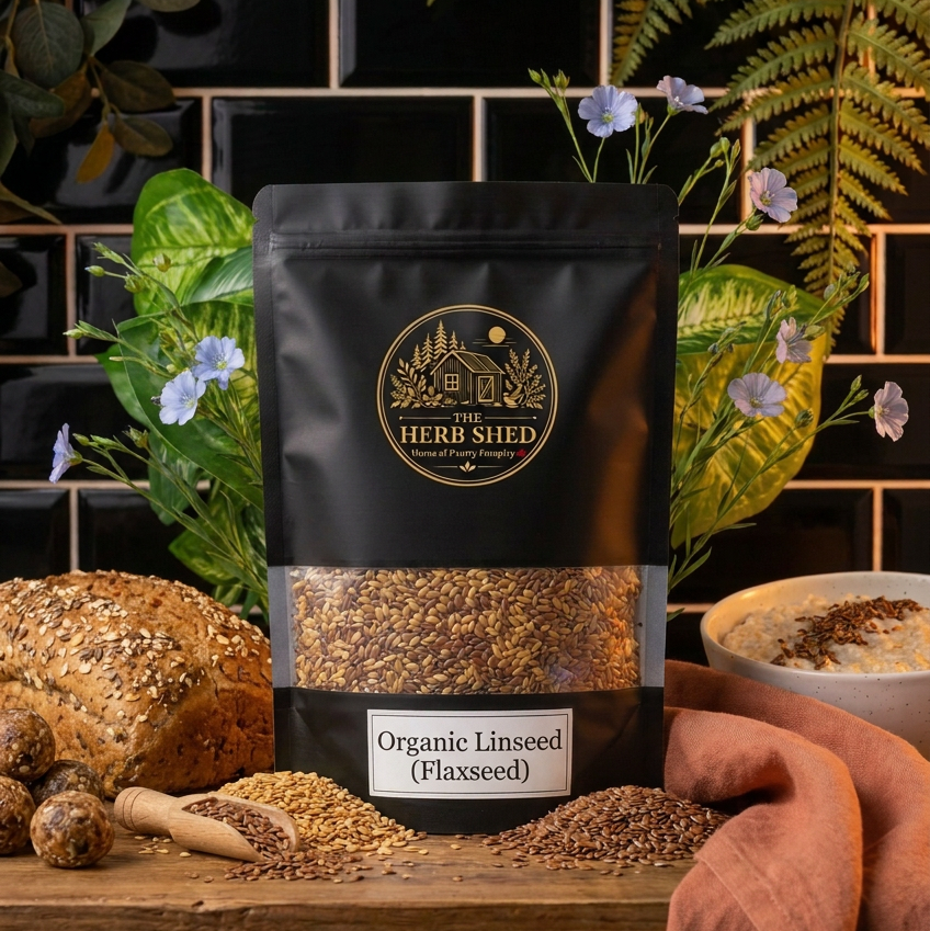 A black stand-up pouch of The Herb Shed Organic Linseed (Flaxseed) sits on a wooden surface with loose brown linseeds, a wooden scoop, seeded bread and green foliage, against a black tiled background with soft terracotta fabric.