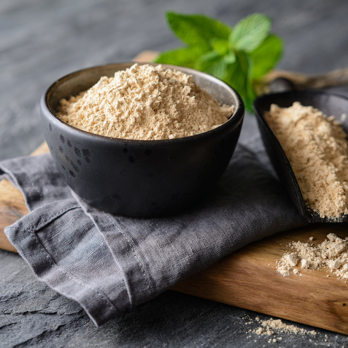 Organic maca powder fills a dark bowl with a wooden scoop and loose powder nearby, showing its finely ground texture and natural beige colouring.