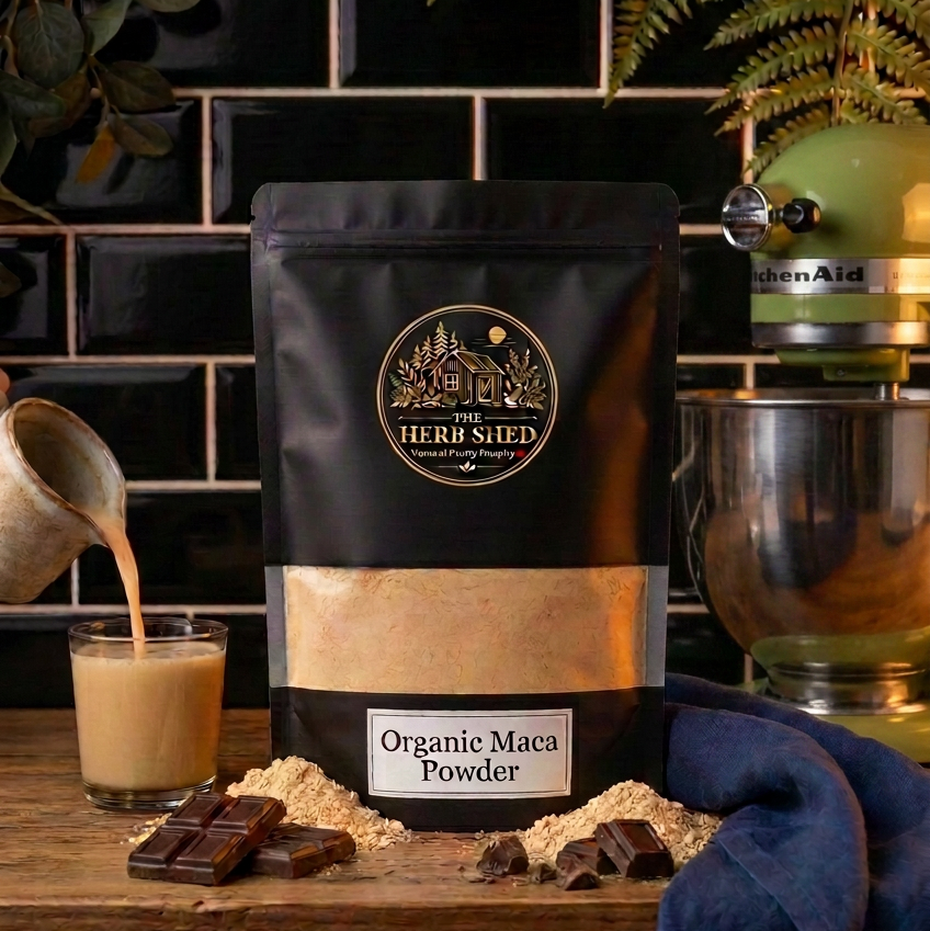 A black stand-up pouch of The Herb Shed Organic Maca Powder sits on a wooden kitchen surface with loose maca powder, a prepared maca drink and pieces of chocolate, with a stand mixer, fern and black tiled background.