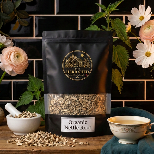 A black stand-up pouch of The Herb Shed Organic Nettle Root sits on a wooden surface with loose dried nettle root, a small white bowl, a teacup, green nettle leaves and soft pink and white flowers, against a black tiled background.