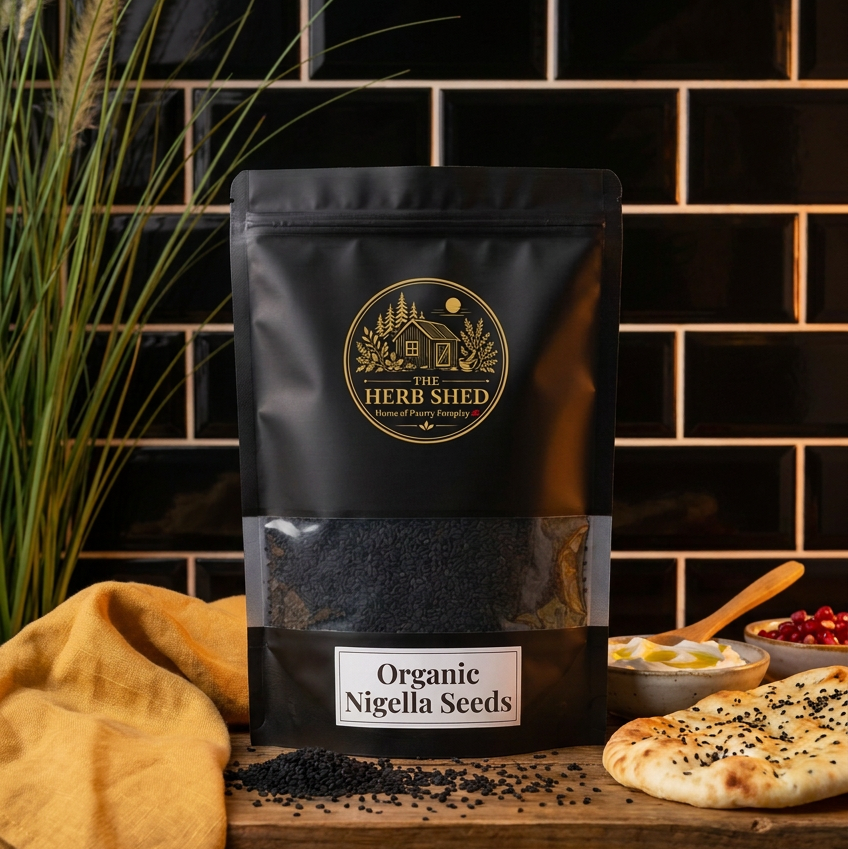 A black stand-up pouch of The Herb Shed Organic Nigella Seeds sits on a wooden surface with loose black nigella seeds and flatbread topped with seeds, surrounded by tall green foliage and warm mustard fabric against a black tiled background.