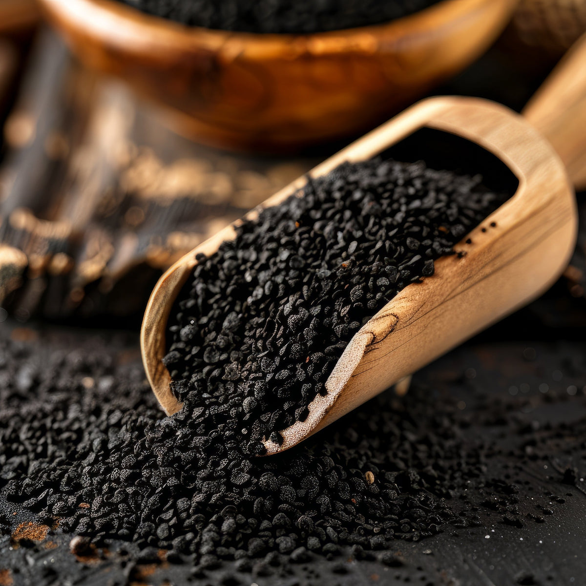 Organic nigella seeds spill from a wooden scoop onto a dark surface, showing their tiny irregular shape, textured surface and deep black colour.