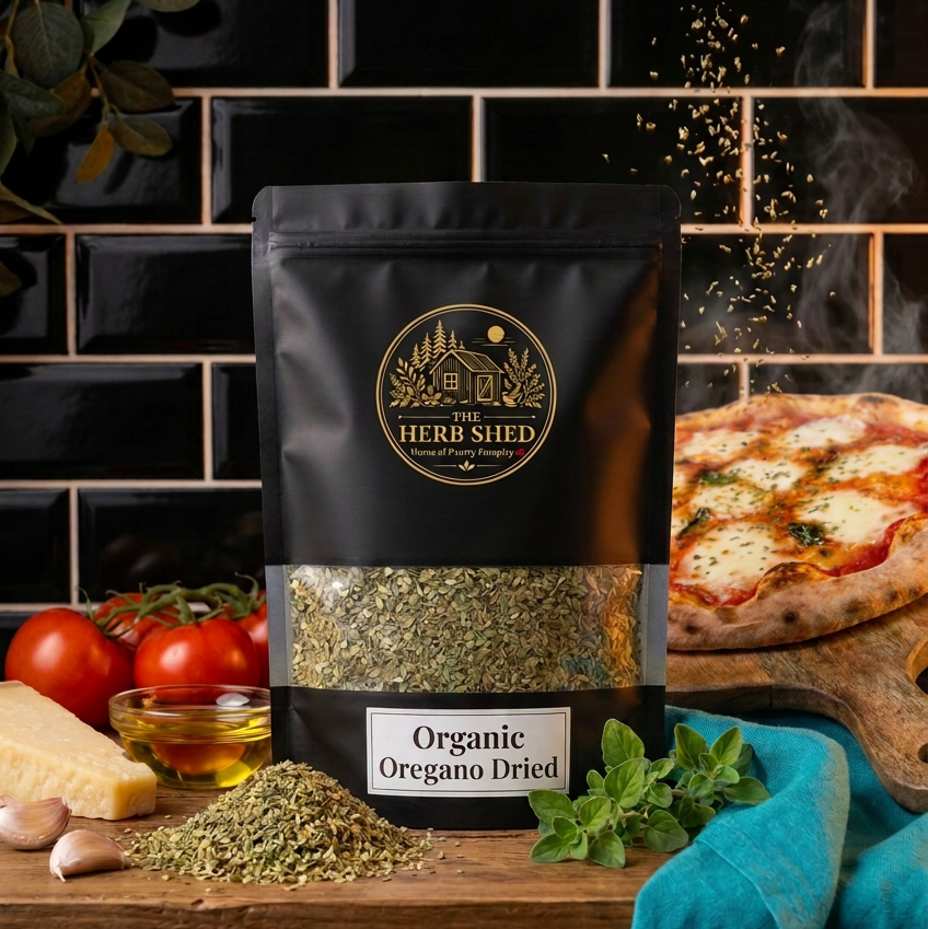 A black stand-up pouch of The Herb Shed Organic Oregano Dried sits on a wooden surface with loose dried oregano, fresh oregano, tomatoes, garlic and olive oil, with a freshly baked pizza against a black tiled kitchen background.
