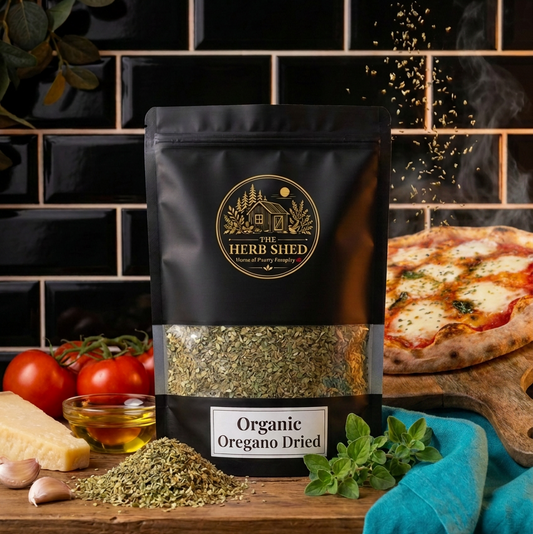 A black stand-up pouch of The Herb Shed Organic Oregano Dried sits on a wooden surface with loose dried oregano, fresh oregano, tomatoes, garlic and olive oil, with a freshly baked pizza against a black tiled kitchen background.