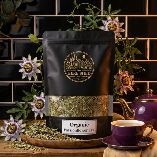 A black stand-up pouch of The Herb Shed Organic Passionflower Tea sits on a wooden surface with loose dried passionflower, purple and white passionflowers and a purple teacup, against a black tiled background.