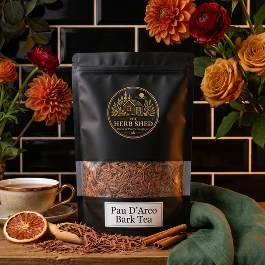 A black stand-up pouch of The Herb Shed Pau D'Arco Bark Tea sits on a wooden surface with loose dried bark, dried citrus, orange and red flowers and a green cloth, against a black tiled background.