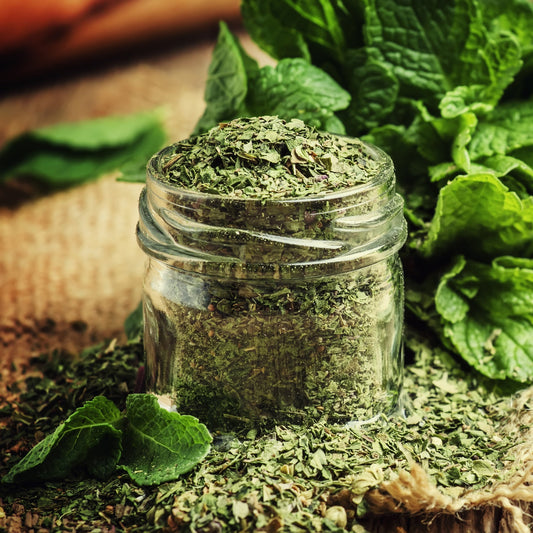A glass jar filled with dried organic peppermint leaf sits among fresh green mint leaves, with loose dried peppermint scattered around the jar.