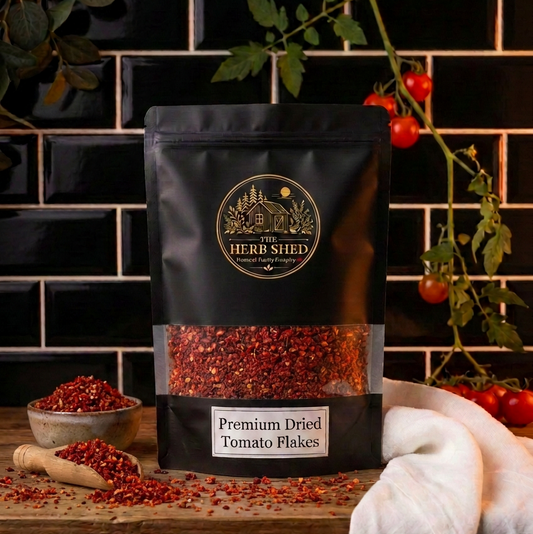A black stand-up pouch of The Herb Shed Premium Dried Tomato Flakes sits on a wooden surface with loose tomato flakes, fresh tomatoes, greenery and a white cloth, against a black tiled background.