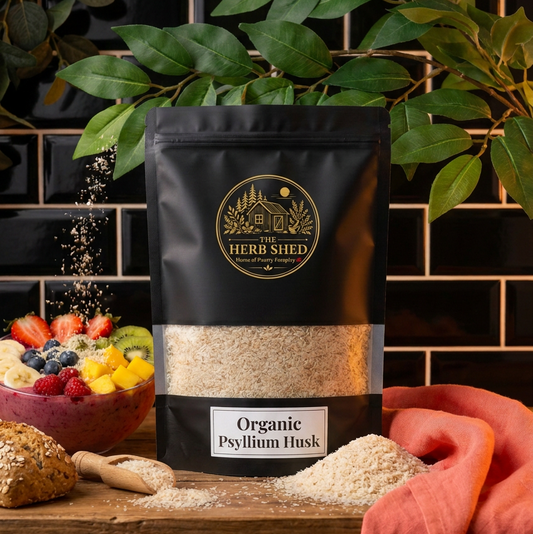 A black stand-up pouch of The Herb Shed Organic Psyllium Husk sits on a wooden surface with loose psyllium husk, a wooden spoon, bread and a bowl of fresh fruit, surrounded by green foliage and coral fabric against a black tiled kitchen background.
