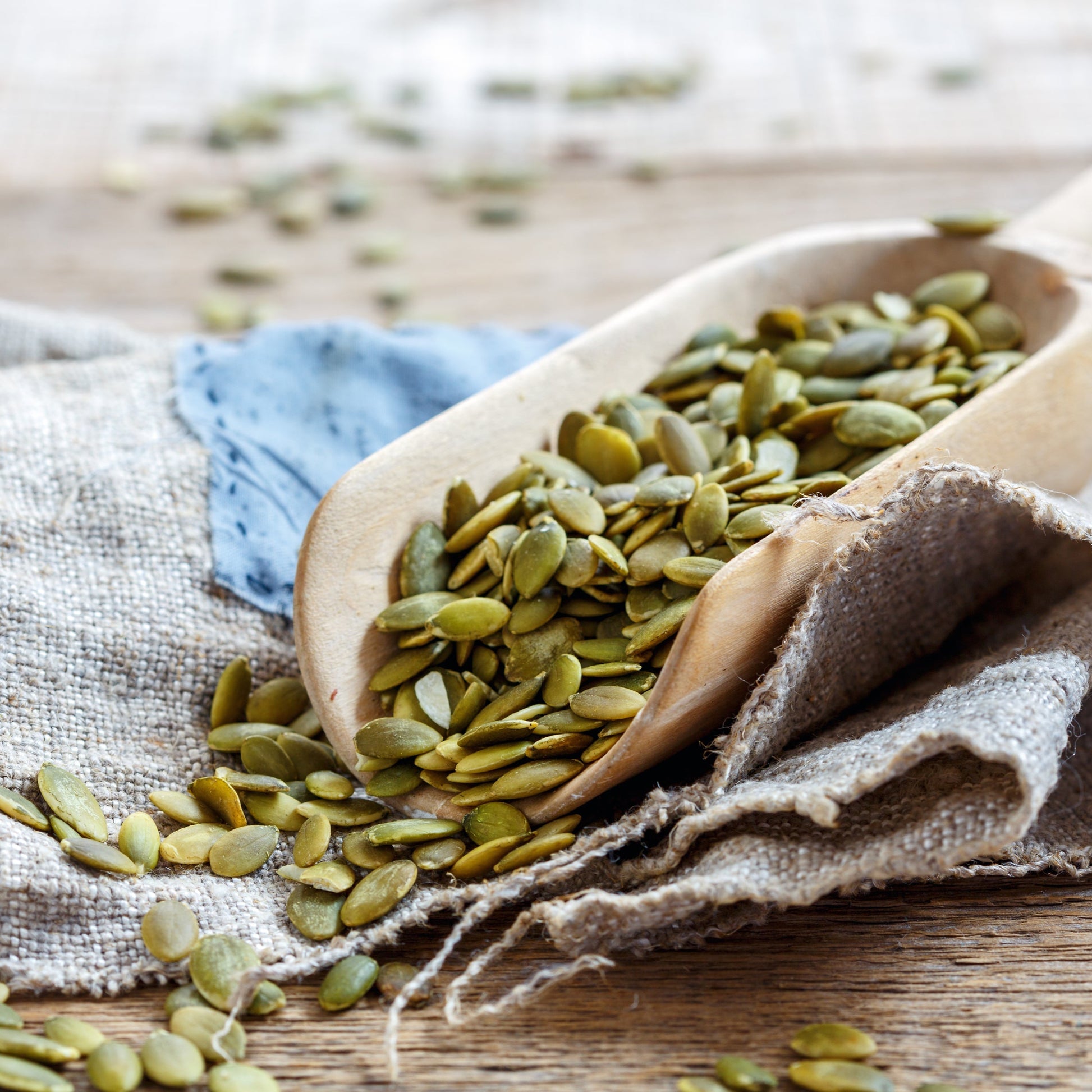 Organic pumpkin seeds fill a white bowl on a light rustic surface, with loose green pumpkin seeds scattered around the bowl and across a soft grey cloth.