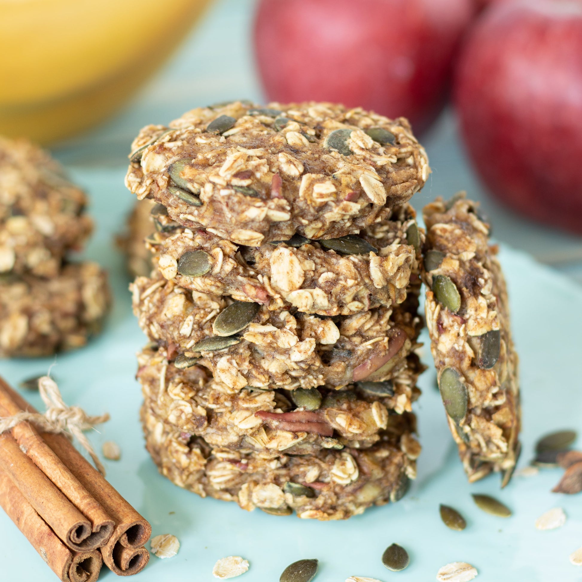 Homemade seed and nut snack bars are stacked on a blue surface with visible pumpkin seeds, nuts and grains, with cinnamon sticks and fresh fruit arranged nearby.