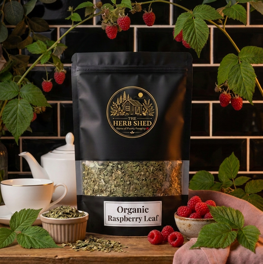 A black stand-up pouch of The Herb Shed Organic Raspberry Leaf sits on a wooden surface with fresh raspberries, raspberry leaves, a white teacup and loose dried leaf, against a black tiled background.