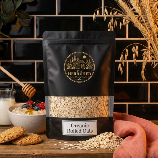 A black stand-up pouch of The Herb Shed Organic Rolled Oats in Wollongong, Australia, sits on a wooden surface with loose rolled oats, oat stems, biscuits, honey and a bowl of oats against a black tiled background.