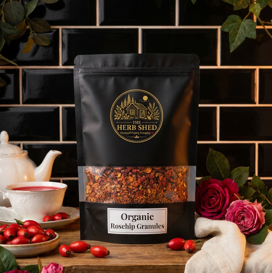 A black stand-up pouch of The Herb Shed Organic Rosehip Granules sits on a wooden surface with loose rosehip granules, red rosehips, red flowers and a white teapot and cup, against a black tiled background.