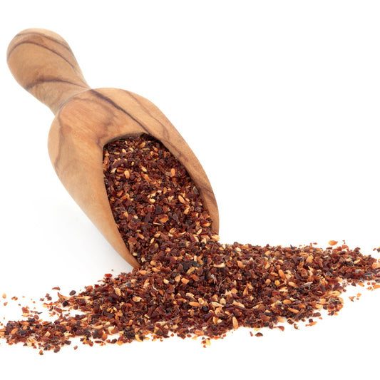 Organic rosehip granules spill from a wooden scoop onto a white surface, showing the coarse red and brown texture of the dried granules.