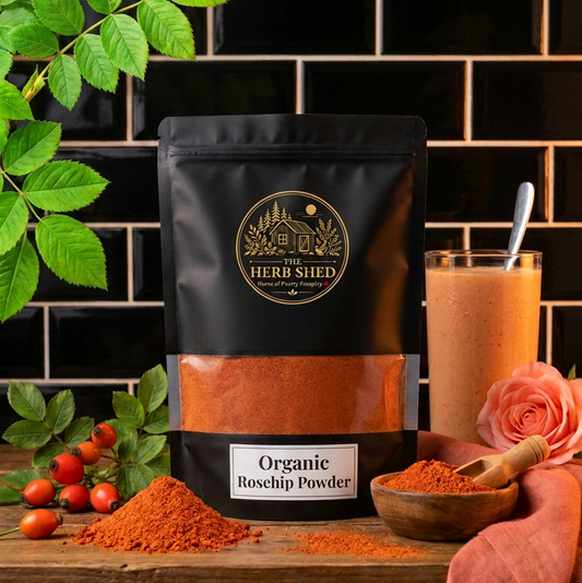 A black stand-up pouch of The Herb Shed Organic Rosehip Powder sits on a wooden surface with loose red-orange rosehip powder, fresh red rosehips, green leaves, a wooden bowl and scoop and a pink rose, against a black tiled background.
