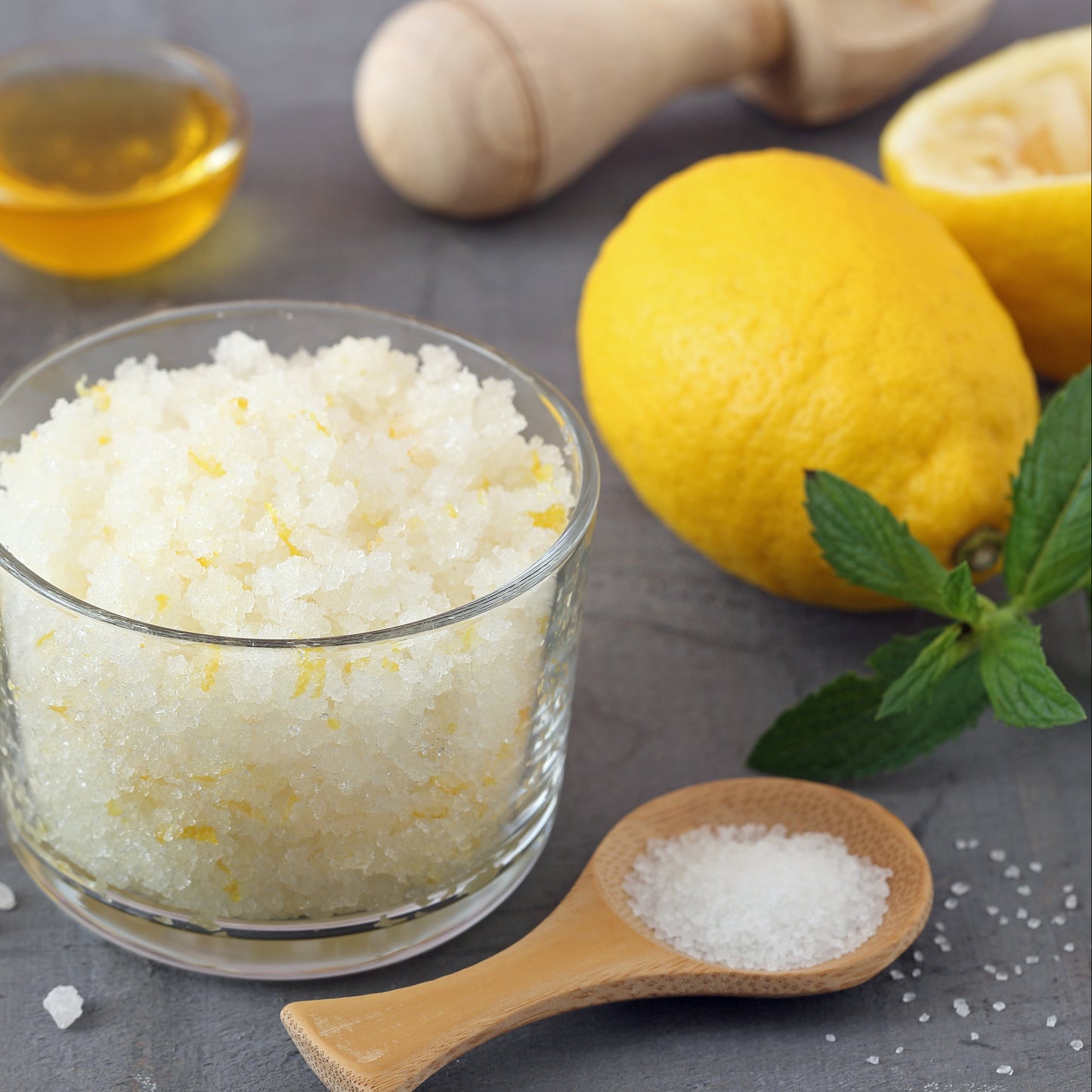 White sea salt flakes sit in a small glass bowl beside fresh lemon, eggs, oil and a wooden spoon of salt on a kitchen surface.