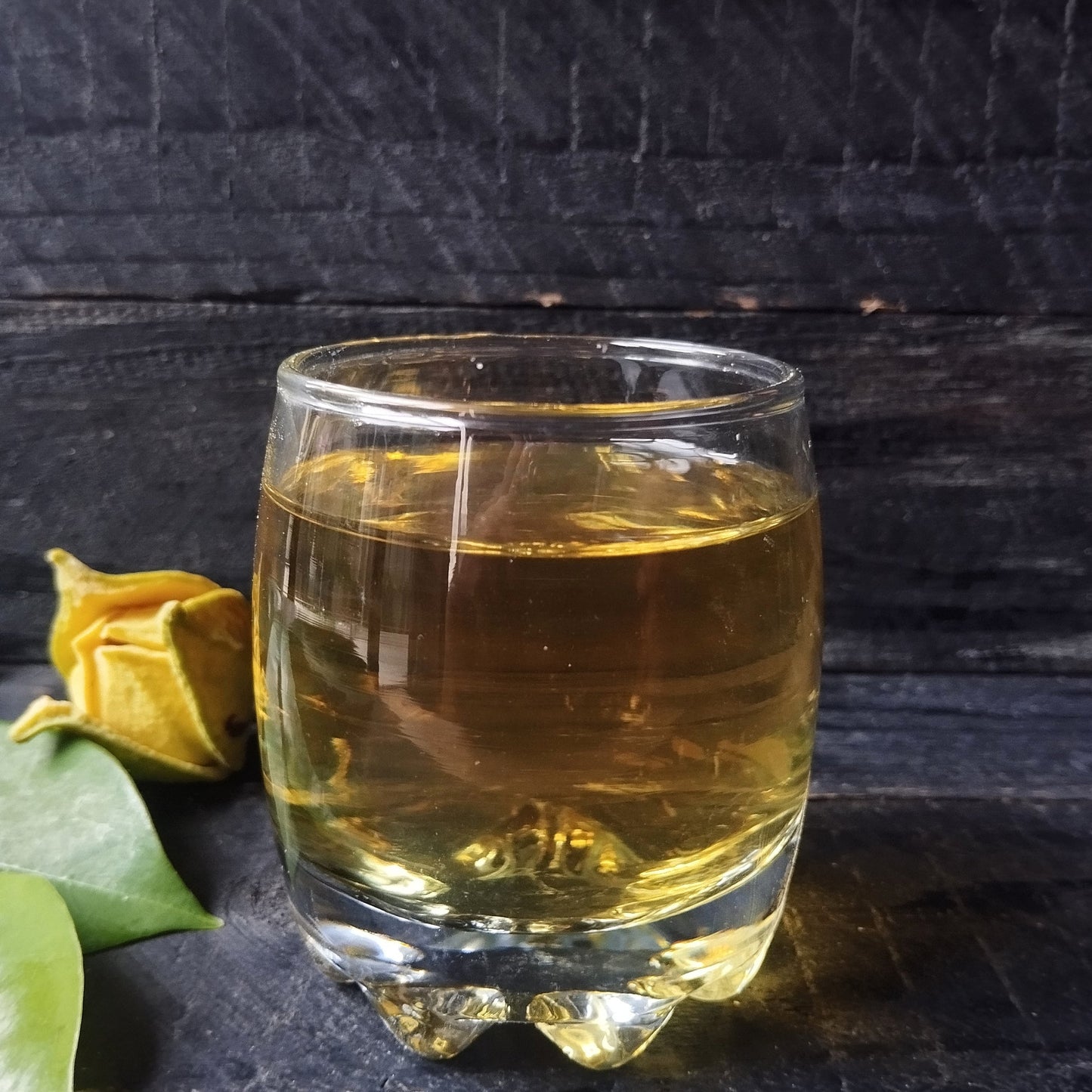 Glass of freshly brewed soursop leaf tea with a warm amber hue, with loose dried leaves beside the cup on a white background from The Herb Shed Wollongong