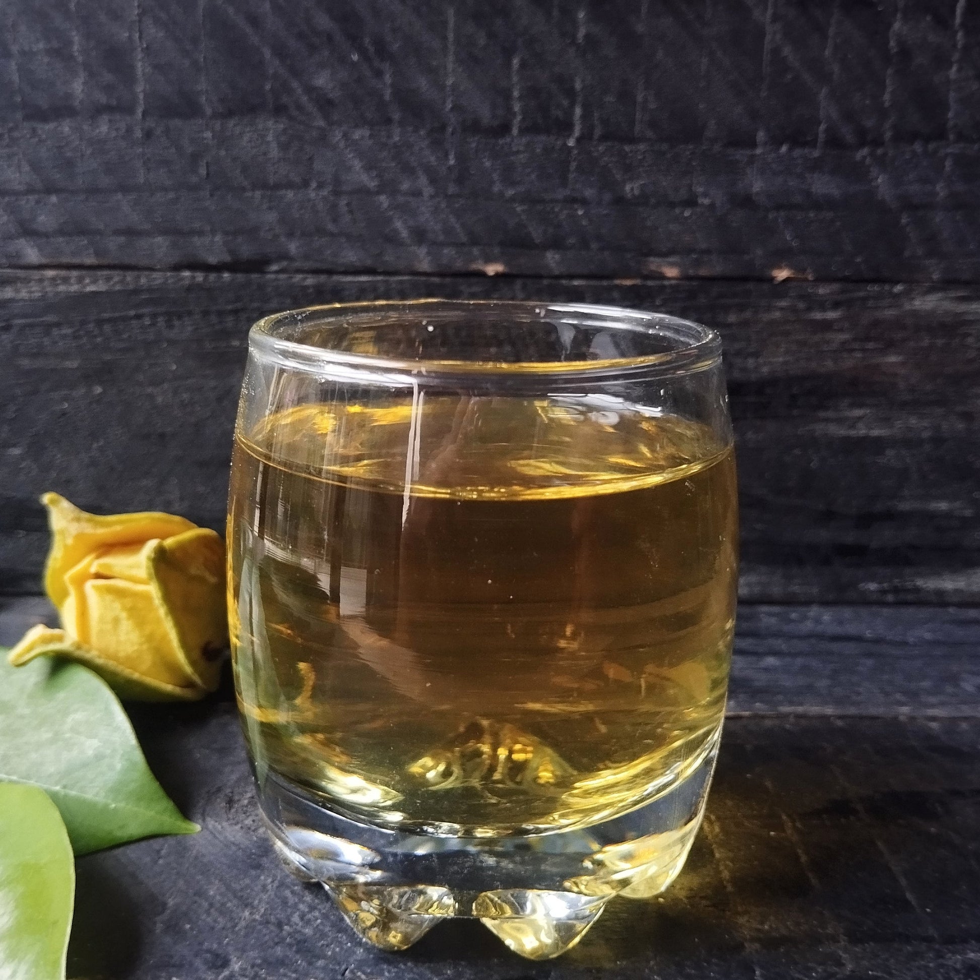Glass of freshly brewed soursop leaf tea with a warm amber hue, with loose dried leaves beside the cup on a white background from The Herb Shed Wollongong