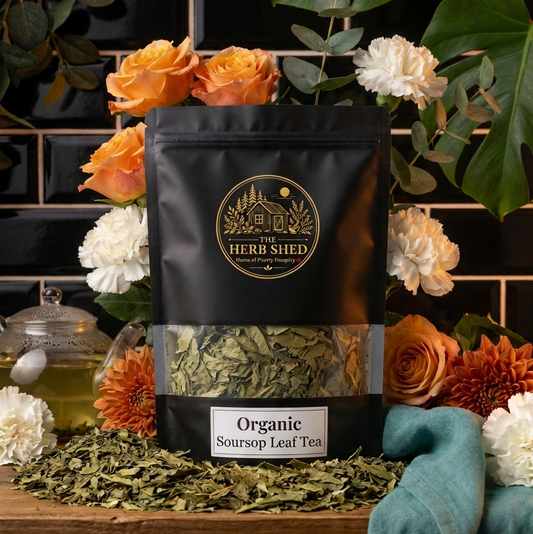 A black stand-up pouch of The Herb Shed Organic Soursop Leaf Tea sits on a wooden surface, surrounded by orange and white flowers, green leaves, and a teal cloth, with a black tiled background.