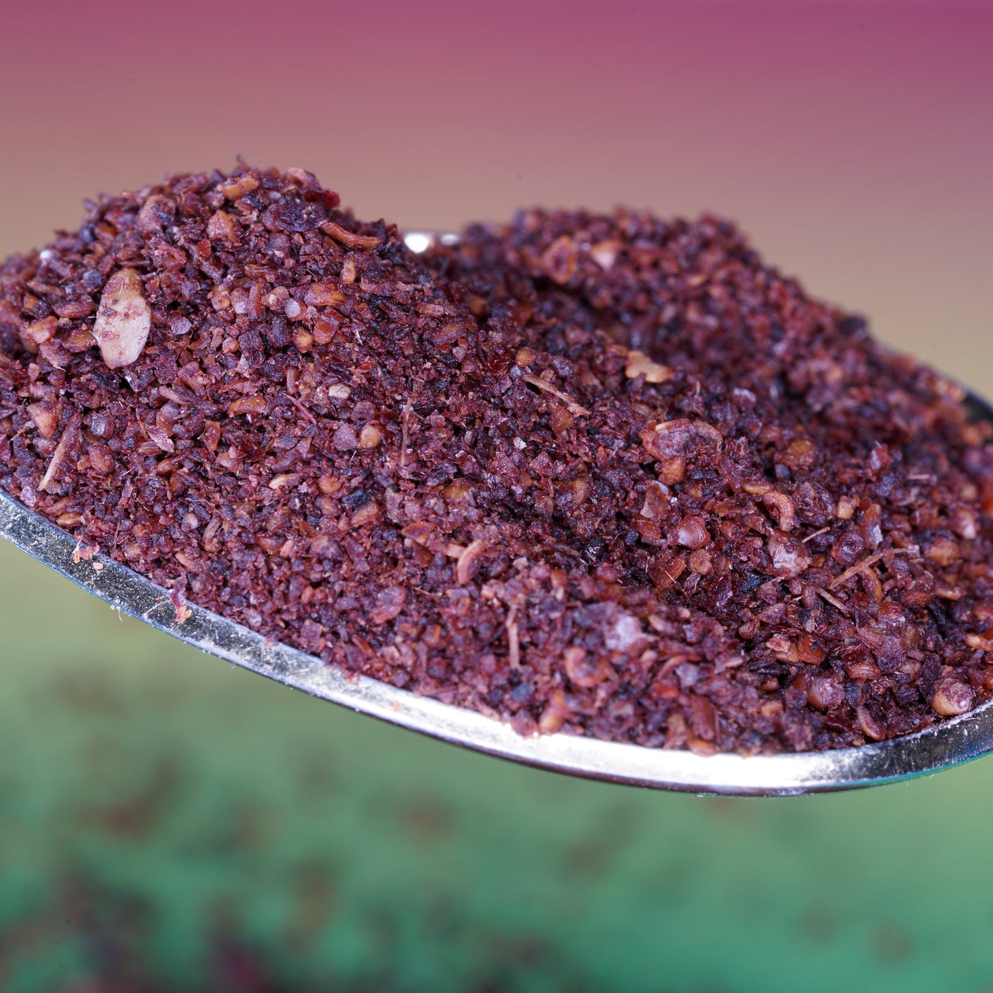 A close-up of organic sumac berry powder piled in a decorative bowl, showing the coarse texture and deep burgundy-red colour of the ground spice.