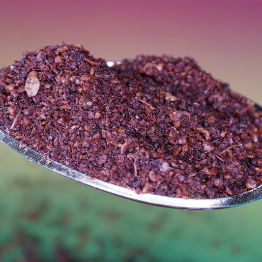 A close-up of organic sumac berry powder piled in a decorative bowl, showing the coarse texture and deep burgundy-red colour of the ground spice.