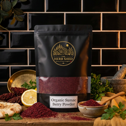 A black stand-up pouch of The Herb Shed Organic Sumac Berry Powder sits on a wooden surface with loose deep red sumac powder, lemon, fresh herbs, spices and rustic kitchenware, against a black tiled kitchen background.