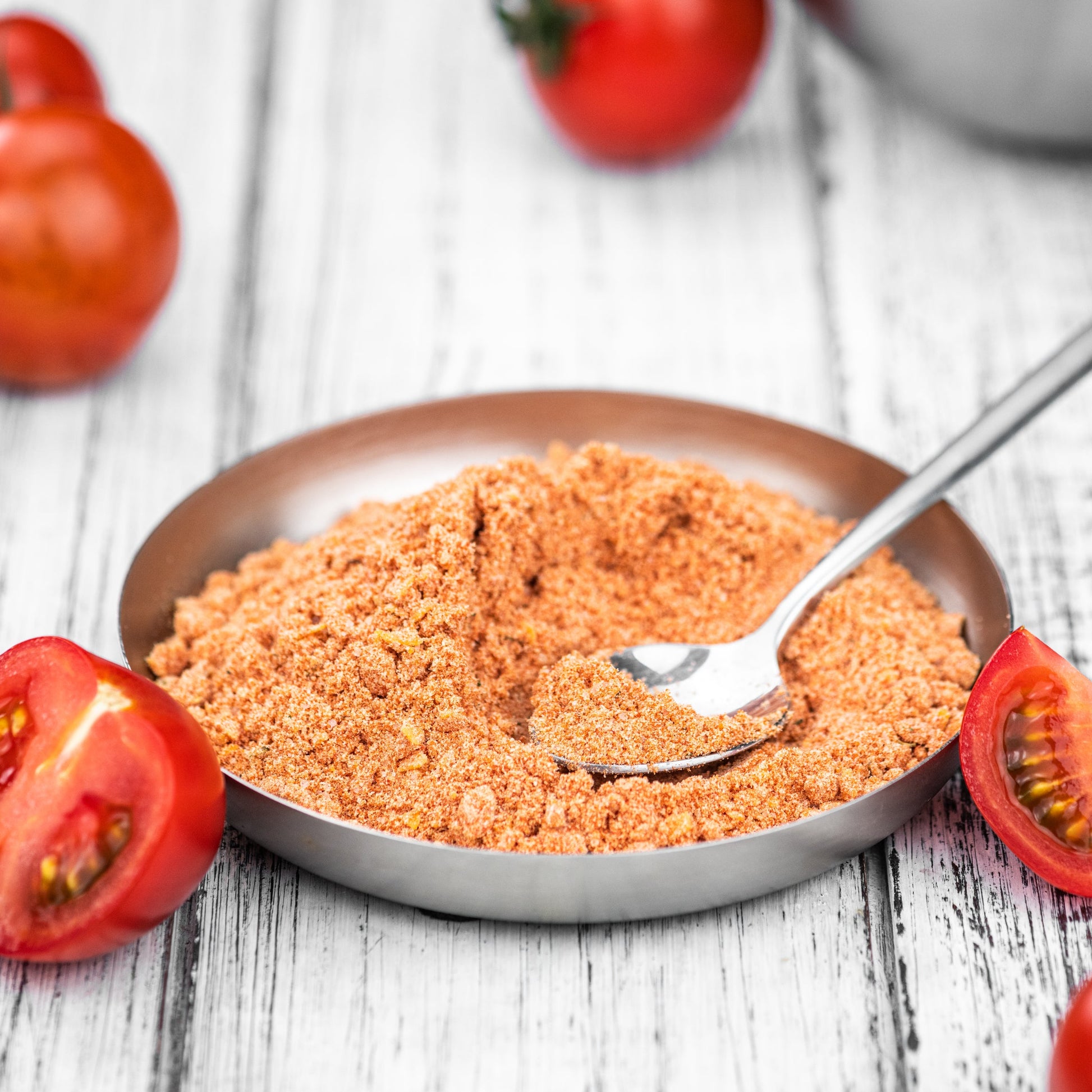 Organic tomato powder sits in a shallow bowl with a small spoon, surrounded by fresh red tomatoes on a light timber surface.