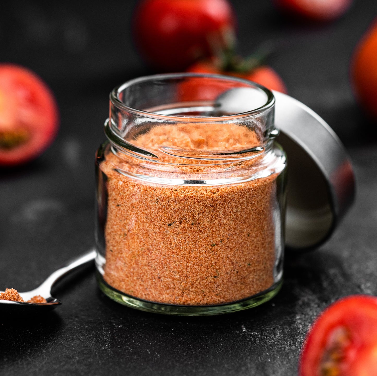 Organic tomato powder fills a small glass jar on a dark surface surrounded by fresh red tomatoes, showing the fine red-orange texture of the powder.