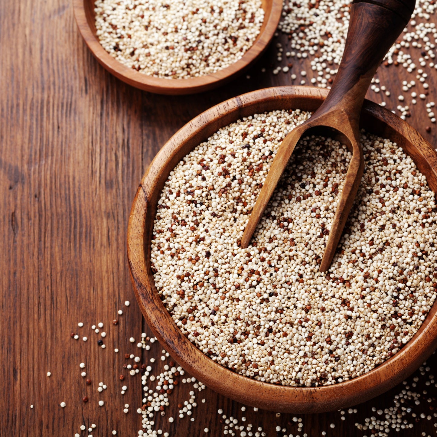Organic tri colour quinoa fills rustic wooden bowls with a wooden scoop, showing the mixture of pale, red and dark quinoa grains on a timber surface.