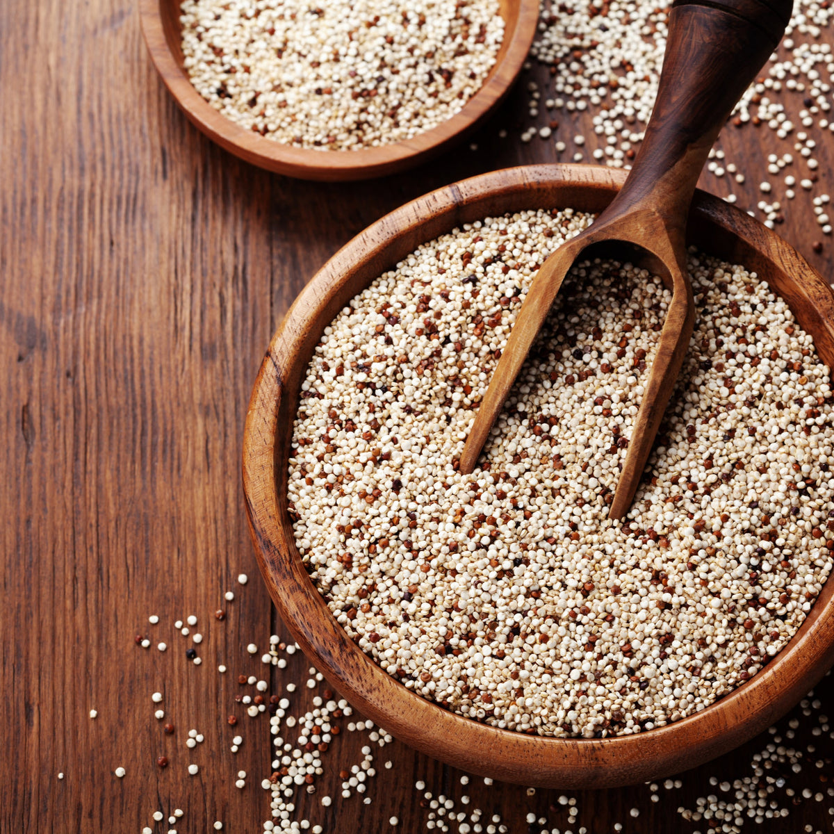 Organic tri colour quinoa fills rustic wooden bowls with a wooden scoop, showing the mixture of pale, red and dark quinoa grains on a timber surface.