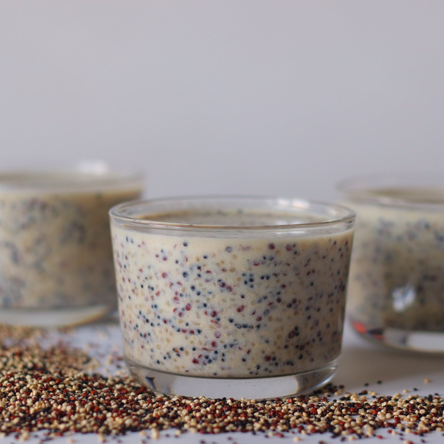 Two glass cups of prepared tri colour quinoa sit on a bed of loose quinoa, showing a creamy serving idea using the mixed quinoa grains.