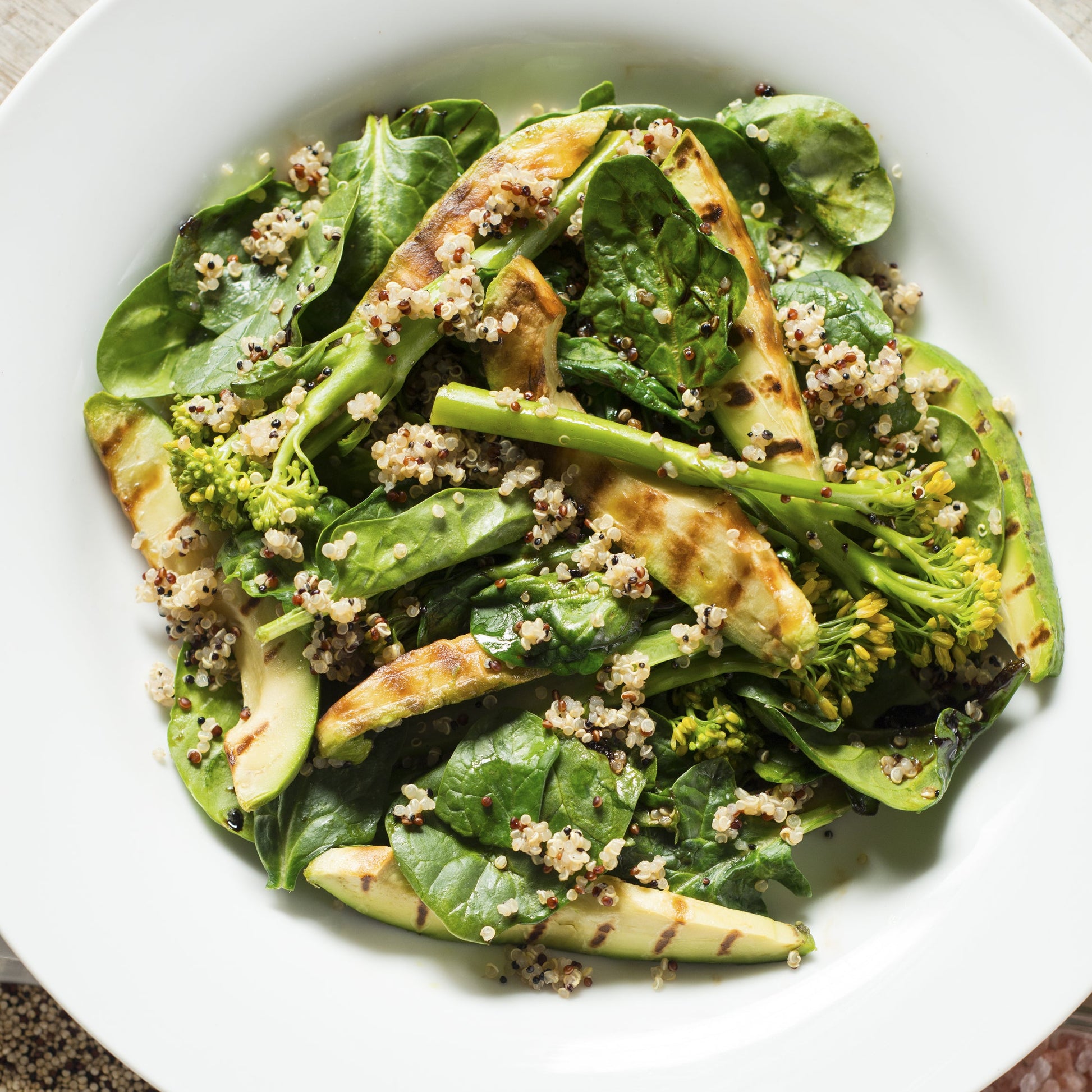 A fresh salad made with organic tri colour quinoa is served with green vegetables and sliced avocado on a white plate.