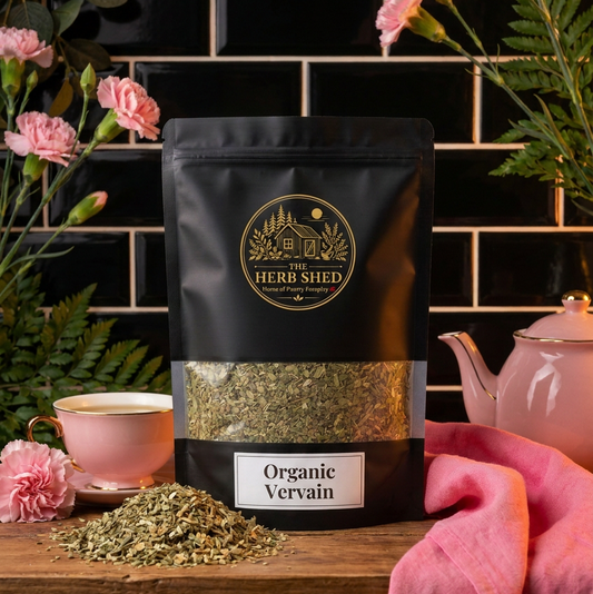 A black stand-up pouch of The Herb Shed Organic Vervain tea with a clear window showing the green herb inside, styled on a wooden surface with pink flowers, a pink teapot, and a pink cup against a black tiled background.