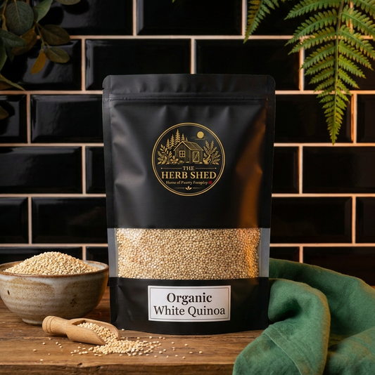 A stand-up pouch of The Herb Shed Organic White Quinoa is displayed on a rustic wooden table against a black subway tile wall, styled with a bowl of loose grains, a wooden spoon, and a green cloth.