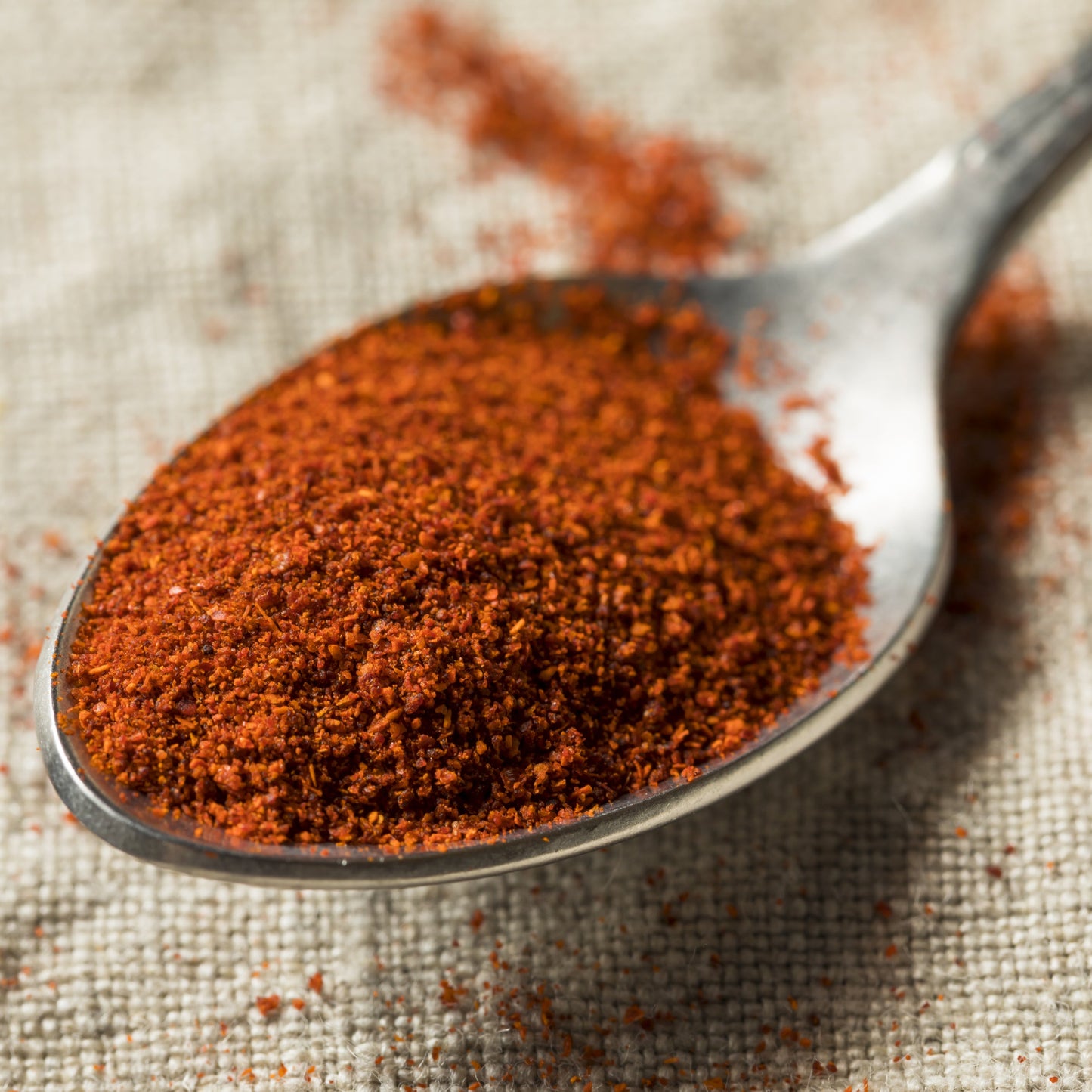 Finely ground smoked paprika fills a metal spoon with loose paprika scattered around it, showing the fine texture and deep red-orange colour of the spice.