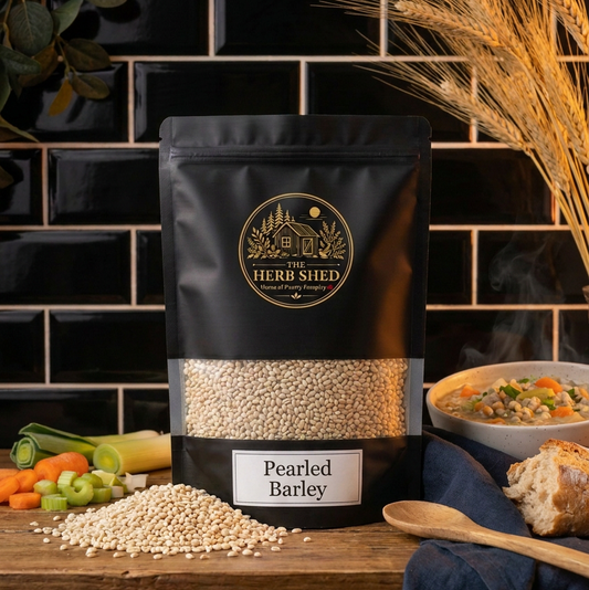 A black stand-up pouch of The Herb Shed Pearled Barley in Wollongong, Australia, sits on a wooden surface with loose barley grains, wheat stems, vegetables, bread and a bowl of hearty barley soup against a black tiled background.