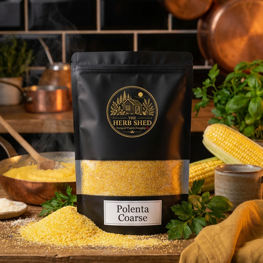 A black stand-up pouch of The Herb Shed Polenta Coarse sits on a wooden surface with loose golden polenta, fresh corn, green herbs, wooden kitchen utensils and warm-toned fabric, against a black tiled kitchen background.