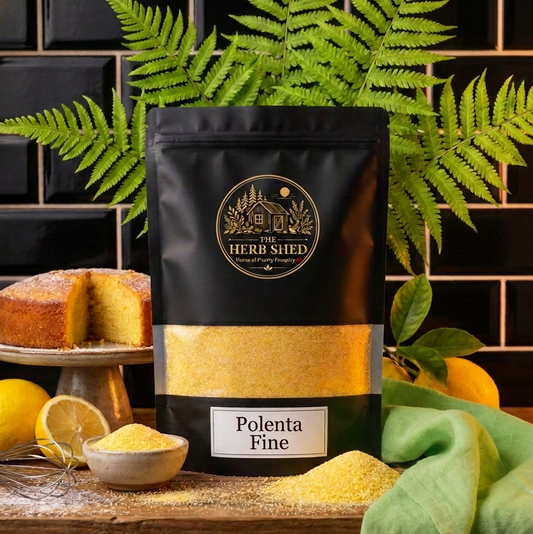 A black stand-up pouch of The Herb Shed Polenta Fine sits on a wooden surface with loose golden polenta, a small bowl, fresh lemons, green foliage and slices of cornbread, with a black tiled background and soft green fabric.