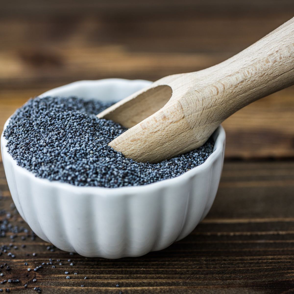 Black poppy seeds fill a small white bowl with a wooden scoop resting across the top, showing the tiny dark seeds against a rustic timber background.