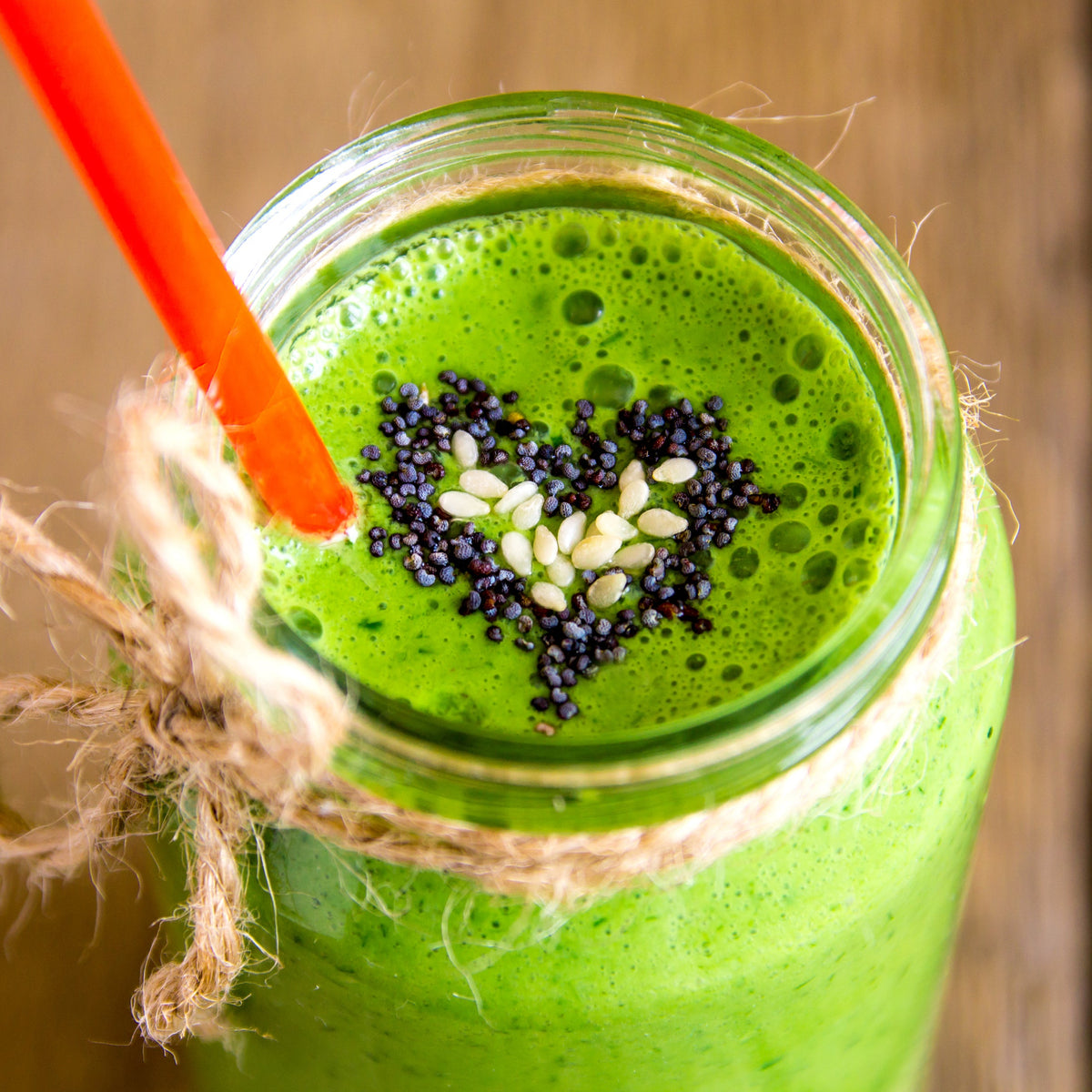 A bright green smoothie in a glass is topped with black poppy seeds, with a red drinking straw and fresh ingredients arranged around the glass.