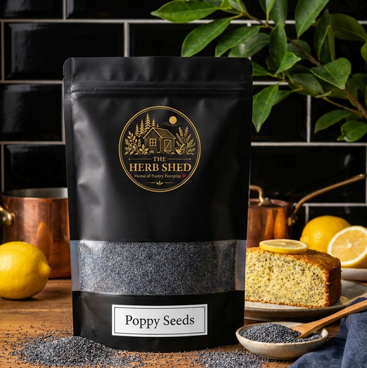 A black stand-up pouch of The Herb Shed Poppy Seeds sits on a wooden kitchen surface with loose black poppy seeds, a wooden spoon, fresh lemons and slices of lemon poppy seed cake, against a black tiled background with green foliage.