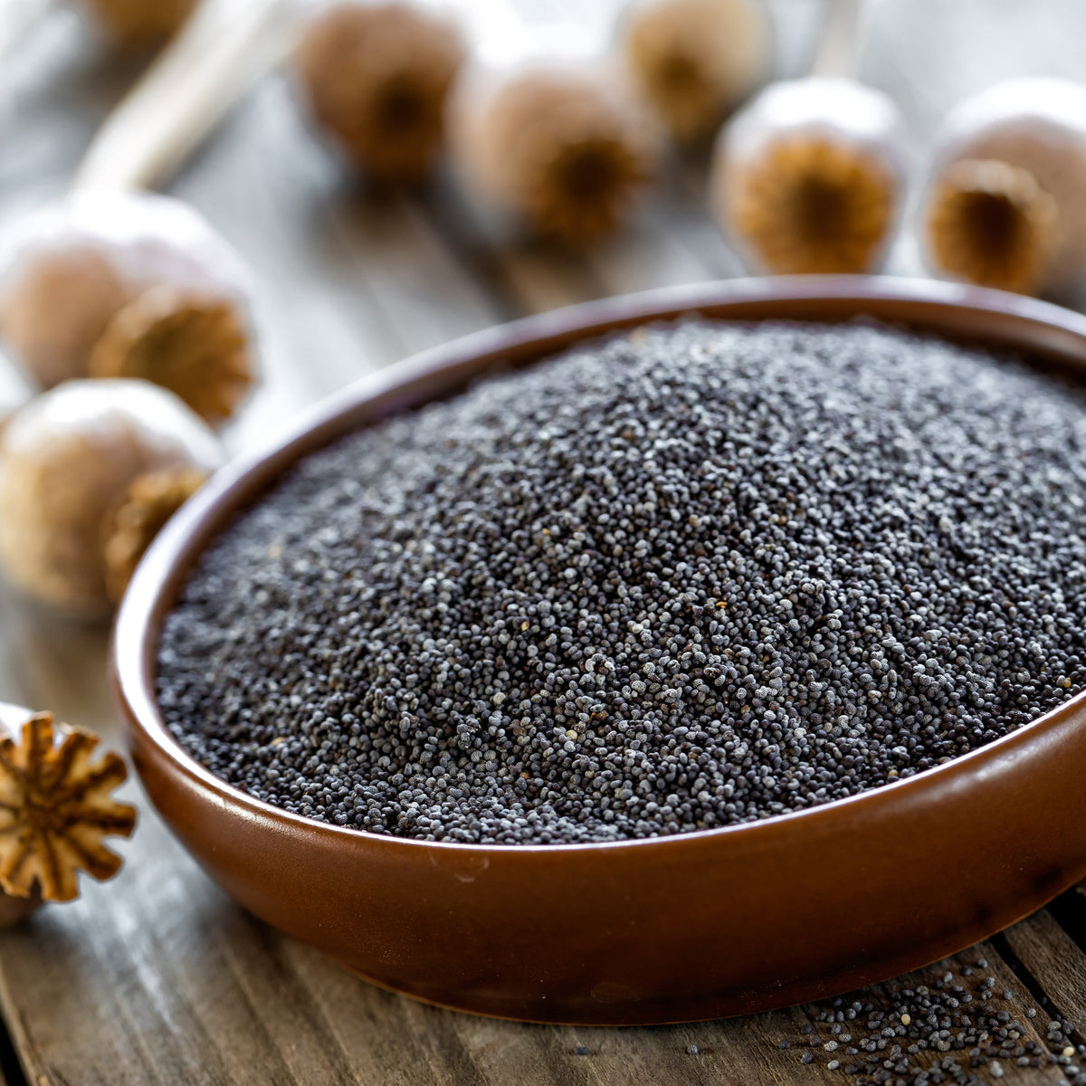 Black poppy seeds fill a round wooden bowl on a rustic surface, showing the tiny dark seeds and their fine granular texture.