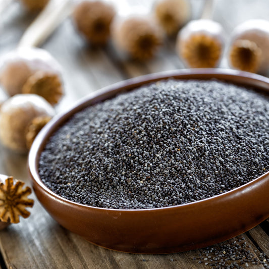 Black poppy seeds fill a round wooden bowl on a rustic surface, showing the tiny dark seeds and their fine granular texture.