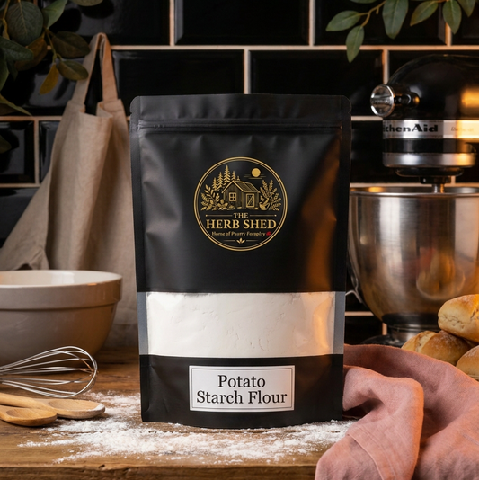 A black stand-up pouch of The Herb Shed Potato Starch Flour sits on a flour-dusted kitchen bench with baking utensils, mixing bowls and a stand mixer, against a black tiled kitchen background with soft pink fabric.