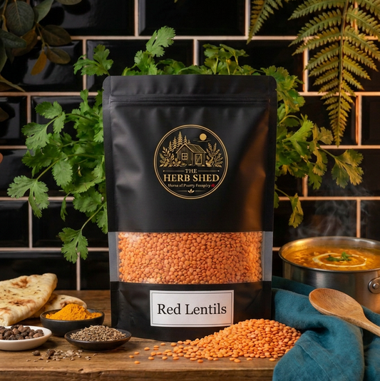 A black stand-up pouch of The Herb Shed Red Lentils in Wollongong, Australia, sits on a wooden surface with loose red lentils, herbs, spices, flatbread and a bowl of lentil curry against a black tiled background.