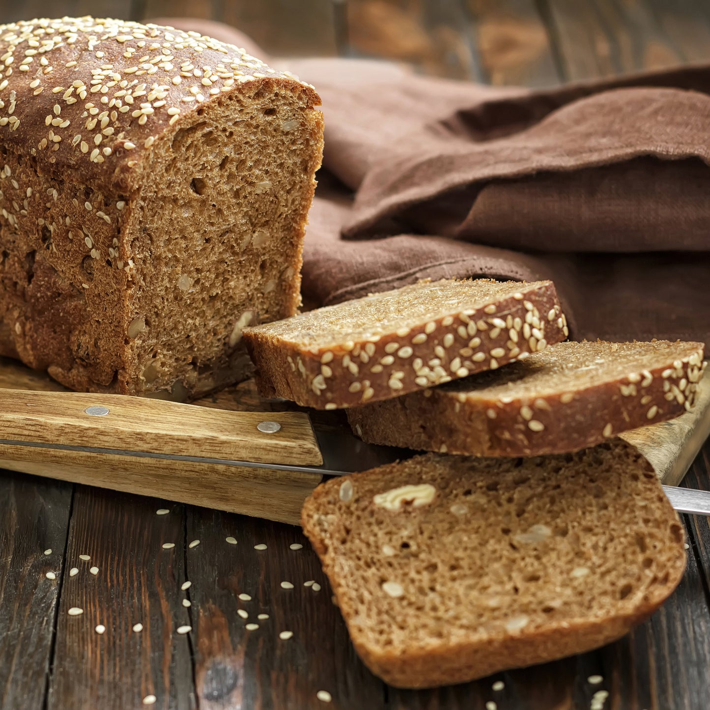 Slices of brown bread coated with sesame seeds sit on a wooden board beside a loaf, showing a baking use for The Herb Shed Roasted Sesame Seeds.