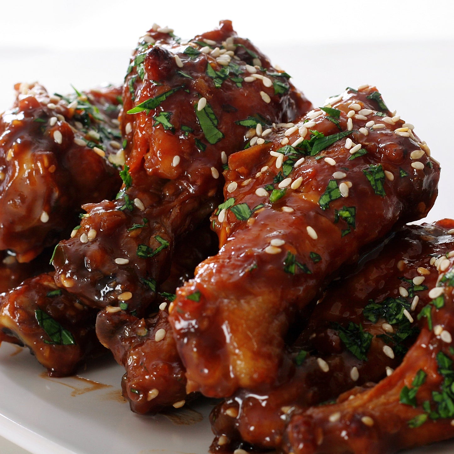 Pieces of glazed chicken coated in a rich red-brown sauce are sprinkled with roasted sesame seeds and fresh green herbs on a white plate.
