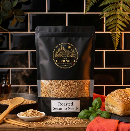 A black stand-up pouch of The Herb Shed Roasted Sesame Seeds sits on a wooden surface with loose sesame seeds, wooden spoons, a bread roll, fresh green herbs and red fabric, against a black tiled kitchen background.